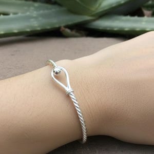 May include: A silver-colored bracelet with a twisted rope design. The bracelet features a teardrop-shaped clasp with a small ball at the top. The bracelet is worn on a person's wrist.