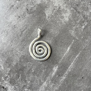 Könnte beinhalten: Silberfarbener Spiralanhänger mit Schlaufe für eine Kette. Der Anhänger hat ein schlichtes, elegantes Design mit glatter, polierter Oberfläche. Die Spiralform ist ein klassisches Symbol.