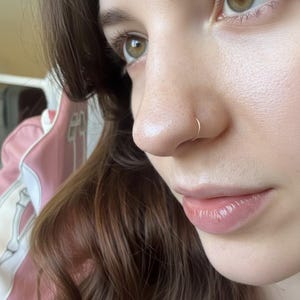 Könnte beinhalten: Nahaufnahme einer Nase mit einem kleinen, dünnen, goldenen Nasenring. Die Haut ist hell mit einem leichten rosa Farbton. Die Person hat braunes Haar und grüne Augen. Der Hintergrund ist verschwommen, mit einer rosa und weißen Jacke sichtbar.