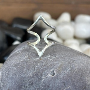 Könnte beinhalten: Ein silberfarbener Ring mit geometrischem Design. Der Ring hat ein Zickzackmuster mit spitzen Enden. Der Ring wird auf einem grauen Stein mit schwarzen und weißen Steinen im Hintergrund präsentiert.