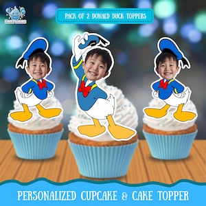 以下が含まれることがあります： 白いフロスティングと青いカップケーキライナーが付いた3つのカップケーキ。各カップケーキには、子供の顔が描かれたドナルドダックをテーマにしたトッパーが付いています。画像には「PACK OF 2 DONALD DUCK TOPPERS」と「PERSONALIZED CUPCAKE & CAKE TOPPER」というテキストが含まれています。