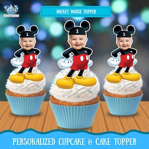 May include: Three cupcakes with white frosting and blue cupcake liners. Each cupcake is topped with a Mickey Mouse-themed cake topper featuring a baby's face. The toppers have black ears, a black shirt, red shorts, yellow shoes, and white gloves. The text "Mickey Mouse Topper" is at the top.