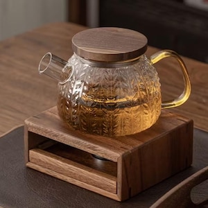 Peut inclure: Une théière en verre transparent avec un couvercle et une poignée en bois, remplie de thé. La théière repose sur une base en bois. Le verre présente un motif texturé. La poignée a une teinte dorée.