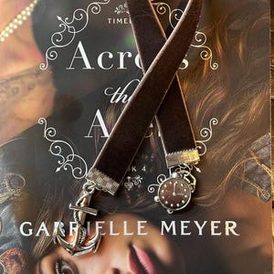 May include: Two brown velvet bookmarks with silver accents. One bookmark features an anchor charm and the other features a clock charm. The bookmarks are laying on a book titled "Across the  Ages" by Gabrielle Meyer.