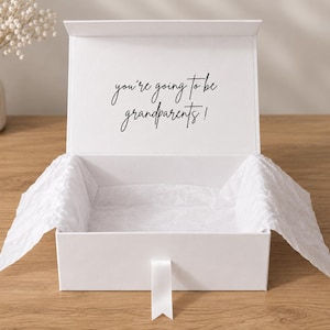 May include: A white rectangular gift box with a hinged lid and a white ribbon pull tab. The interior of the lid features the black printed text "you're going to be grandparents!". The box is lined with white crinkled tissue paper.