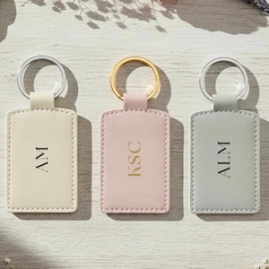 May include: Three rectangular keyrings in cream, pink, and grey, each with a silver or gold ring. The cream keyring has the initials "AM", the pink keyring has "KSC", and the grey keyring has "ALM".