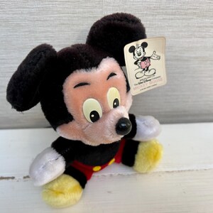 Puede incluir: Un peluche de Mickey Mouse con pelaje negro, cara rosa y pies amarillos. El juguete lleva un atuendo rojo y negro. Una etiqueta con una ilustraci&oacute;n de Mickey Mouse est&aacute; adjunta.