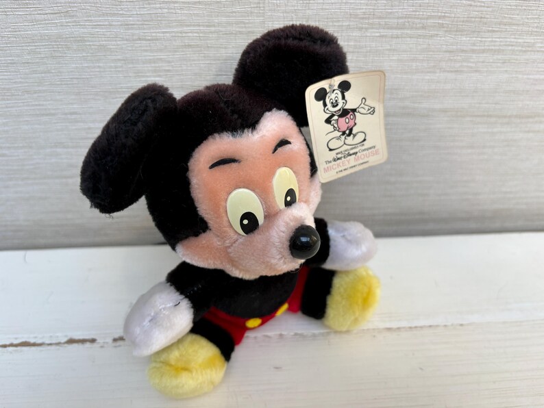 Puede incluir: Un peluche de Mickey Mouse con pelaje negro, cara rosa y pies amarillos. El juguete lleva un atuendo rojo y negro. Una etiqueta con una ilustraci&oacute;n de Mickey Mouse est&aacute; adjunta.
