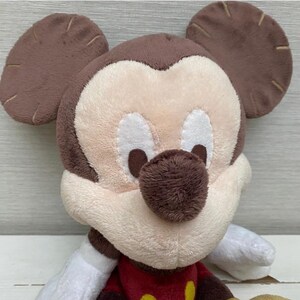May include: A plush Mickey Mouse toy with a brown head and ears, a cream-coloured face, and a brown nose. The toy has white gloves and a red shirt with yellow buttons. The toy is made of soft, plush material.