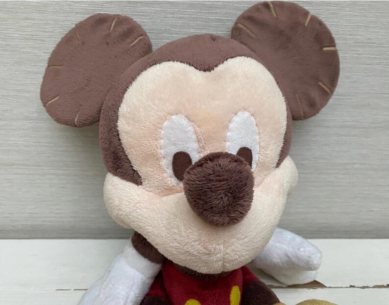 May include: A plush Mickey Mouse toy with a brown head and ears, a cream-coloured face, and a brown nose. The toy has white gloves and a red shirt with yellow buttons. The toy is made of soft, plush material.