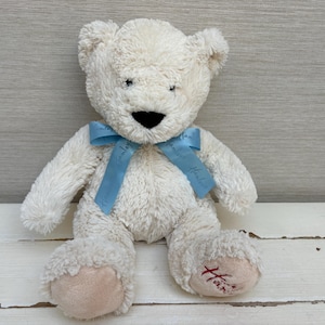 May include: A cream-colored plush teddy bear with soft, shaggy fur, a black stitched nose, and a light blue satin ribbon tied around its neck. The bear features smooth, light tan fabric on its foot pads, with the word Hamleys embroidered in red.