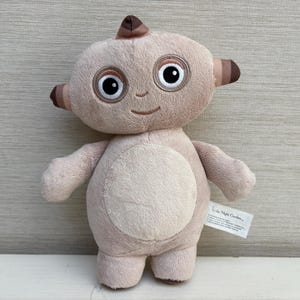 Może przedstawiać: Pluszowa zabawka przedstawiająca postać Igglepiggle z In the Night Garden, wykonana z miękkiego, jasnobrązowego materiału, z okrągłą łatą na brzuchu, dużymi białymi oczami z czarnymi źrenicami i małym brązowym kosmykiem włosów na głowie.