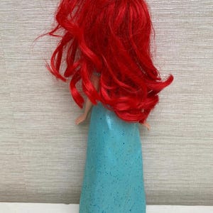 Op de afbeelding: Pop met lang, felrood haar en een turquoise jurk. Het haar van de pop is gestyled in losse golven en de jurk heeft een glitterafwerking. De pop is een verzamelobject, waarschijnlijk een personage uit een populaire animatiefilm.