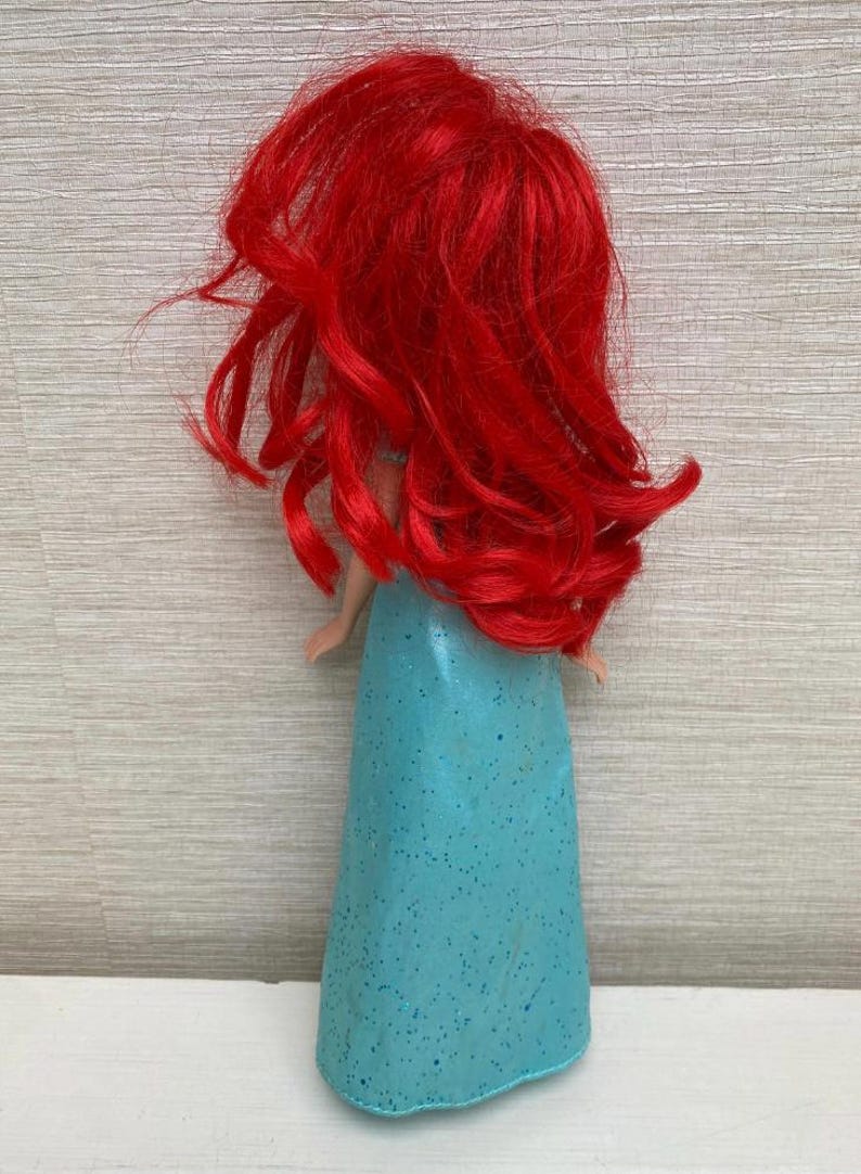 Op de afbeelding: Pop met lang, felrood haar en een turquoise jurk. Het haar van de pop is gestyled in losse golven en de jurk heeft een glitterafwerking. De pop is een verzamelobject, waarschijnlijk een personage uit een populaire animatiefilm.