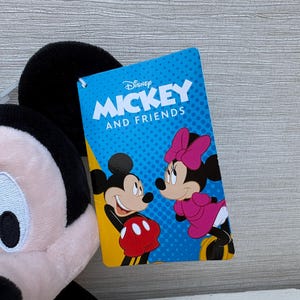 May include: A plush Mickey Mouse toy with a Disney "Mickey and Friends" tag. The tag features Mickey and Minnie Mouse illustrations on a blue background. The plush toy has black ears, a pink face, and a red and white outfit.