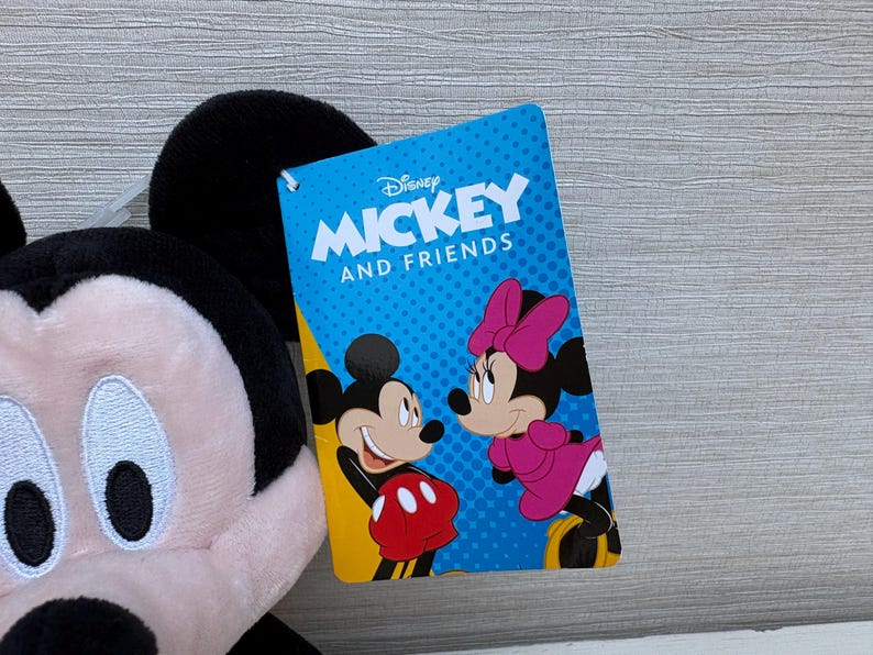 May include: A plush Mickey Mouse toy with a Disney "Mickey and Friends" tag. The tag features Mickey and Minnie Mouse illustrations on a blue background. The plush toy has black ears, a pink face, and a red and white outfit.