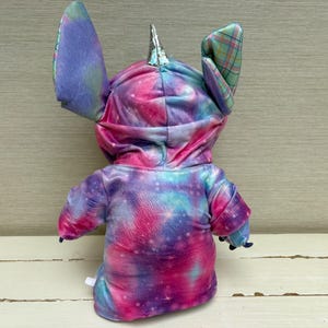 May include: A plush toy in the shape of a character with large ears and a horn. The toy is wearing a hooded sweatshirt with a tie-dye galaxy pattern in shades of pink, purple, and blue. The ears are a mix of tie-dye and plaid.