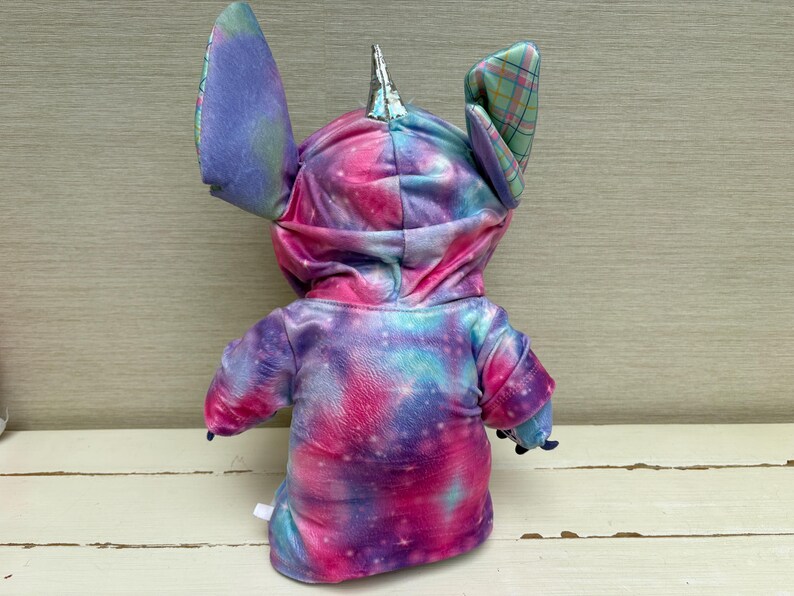 May include: A plush toy in the shape of a character with large ears and a horn. The toy is wearing a hooded sweatshirt with a tie-dye galaxy pattern in shades of pink, purple, and blue. The ears are a mix of tie-dye and plaid.