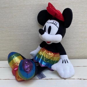May include: A Minnie Mouse soft toy with a red bow, black ears, and a rainbow-coloured skirt and shoes. The doll is sitting on a white surface.