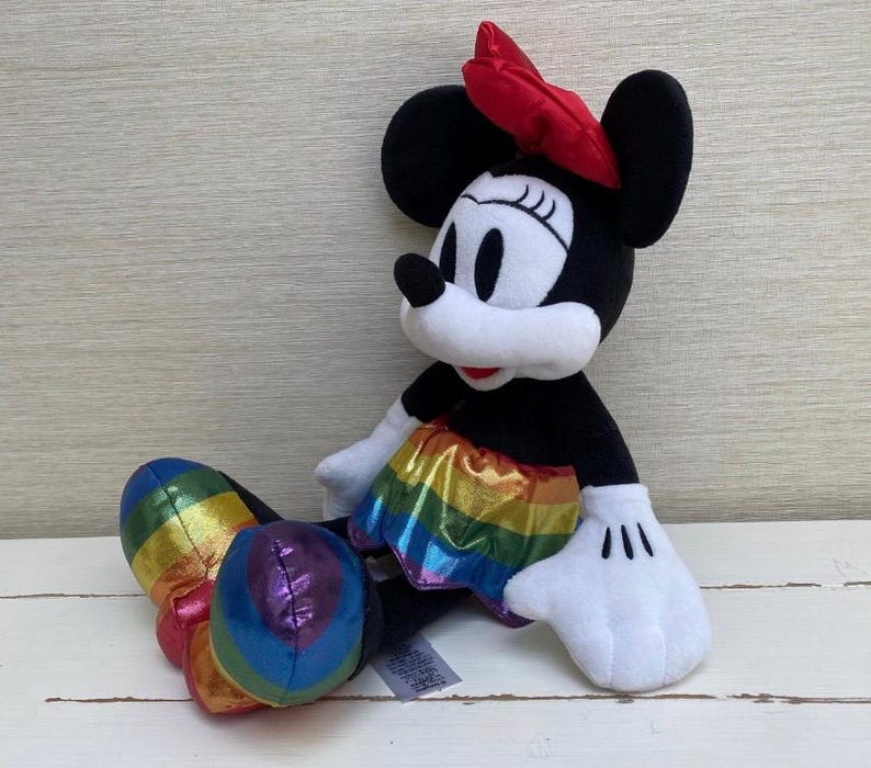 May include: A Minnie Mouse soft toy with a red bow, black ears, and a rainbow-coloured skirt and shoes. The doll is sitting on a white surface.
