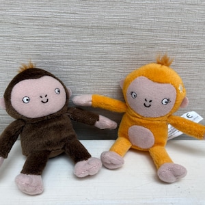May include: Two plush monkey toys, one dark brown and one bright orange, each featuring a light pink face, small ears, and embroidered facial features. The orange monkey has a small star detail on its head, and both have soft limbs and bodies.