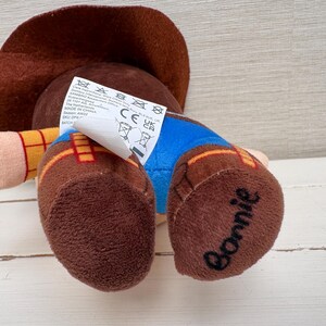 May include: A plush toy of a cowboy character, wearing a brown hat, yellow shirt, blue pants, and brown boots. The word "Bonnie" is written on one of the boots. A tag with care instructions is attached.