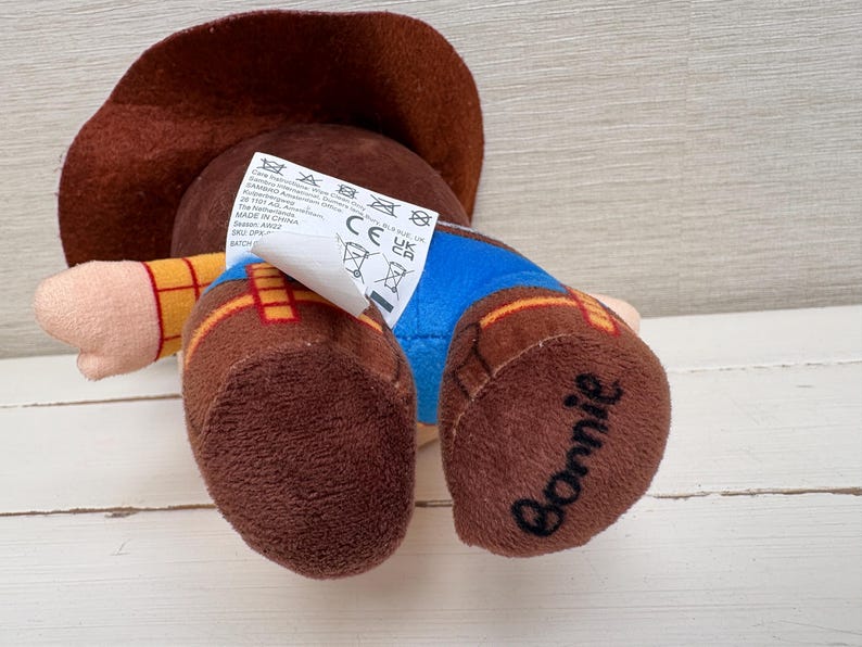 May include: A plush toy of a cowboy character, wearing a brown hat, yellow shirt, blue pants, and brown boots. The word "Bonnie" is written on one of the boots. A tag with care instructions is attached.