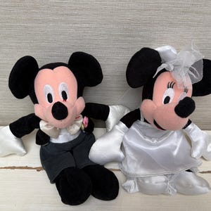 May include: Two plush Mickey and Minnie Mouse dolls dressed for a wedding. Mickey wears a tuxedo with a bow tie and a pink flower. Minnie is in a white wedding dress with a veil. Both have black ears and pink faces.