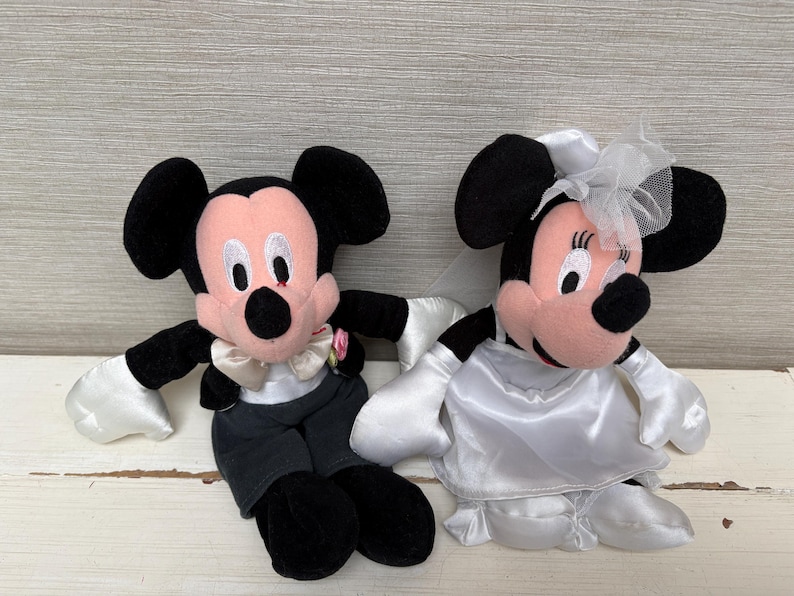 May include: Two plush Mickey and Minnie Mouse dolls dressed for a wedding. Mickey wears a tuxedo with a bow tie and a pink flower. Minnie is in a white wedding dress with a veil. Both have black ears and pink faces.
