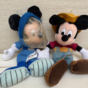 May include: Two Mickey Mouse soft toys. One is dressed in a blue astronaut suit with a clear plastic helmet. The other Mickey Mouse is wearing a red shirt, denim overalls, and a yellow cowboy hat. Both have black ears and white gloves.