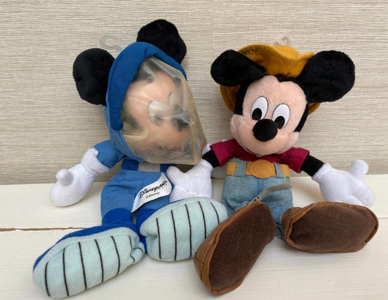May include: Two Mickey Mouse soft toys. One is dressed in a blue astronaut suit with a clear plastic helmet. The other Mickey Mouse is wearing a red shirt, denim overalls, and a yellow cowboy hat. Both have black ears and white gloves.