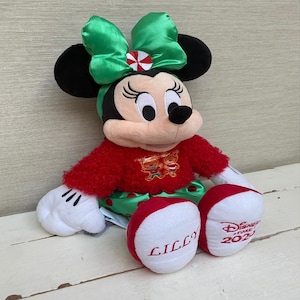 May include: A plush Minnie Mouse doll, featuring a red sweater and green skirt. The doll has a large green bow with a red and white accent. The doll's feet are red and white, with the name "LILLY" embroidered on one foot and "Disney Store 2023" on the other.
