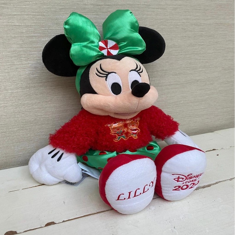 May include: A plush Minnie Mouse doll, featuring a red sweater and green skirt. The doll has a large green bow with a red and white accent. The doll's feet are red and white, with the name "LILLY" embroidered on one foot and "Disney Store 2023" on the other.