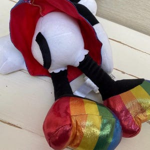 May include: A soft toy featuring a white head and feet, black legs, and rainbow-striped shoes. It has a red cape with a blue lining. The toy is positioned on a light-coloured wooden surface.
