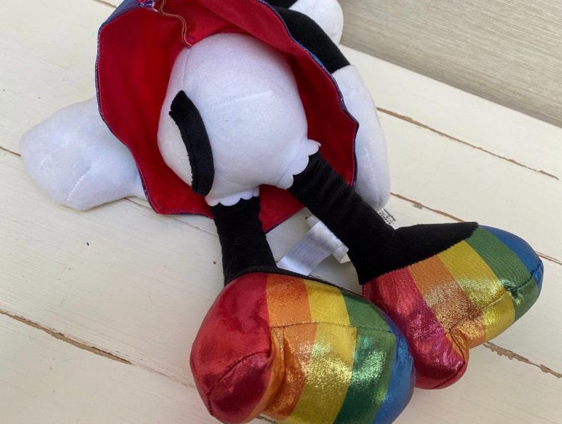 May include: A soft toy featuring a white head and feet, black legs, and rainbow-striped shoes. It has a red cape with a blue lining. The toy is positioned on a light-coloured wooden surface.