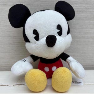 Op de afbeelding: Een pluche Mickey Mouse-speelgoed met een wit gezicht, zwarte oren en een zwarte neus. Mickey draagt een rode broek met twee witte knopen en gele voeten. Het speelgoed heeft witte handschoenen en een zwart-wit gestreepte torso.
