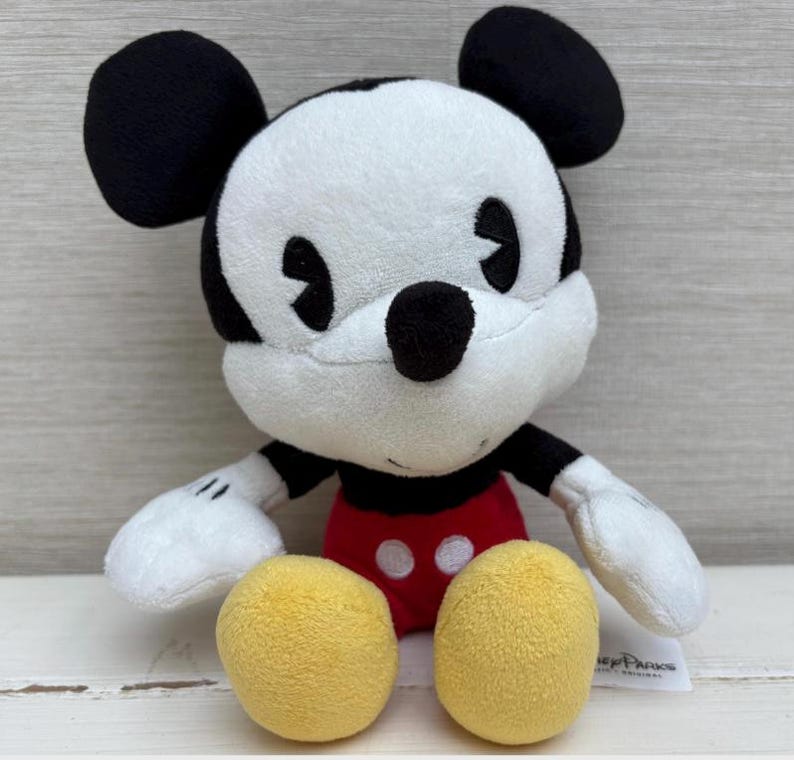 Op de afbeelding: Een pluche Mickey Mouse-speelgoed met een wit gezicht, zwarte oren en een zwarte neus. Mickey draagt een rode broek met twee witte knopen en gele voeten. Het speelgoed heeft witte handschoenen en een zwart-wit gestreepte torso.