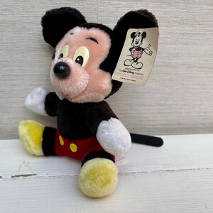 Puede incluir: Un peluche de Mickey Mouse con detalles en negro, rojo, amarillo y blanco. El juguete tiene una cara rosa, orejas negras y una cola negra. Una etiqueta con el logotipo de Mickey Mouse est&aacute; adjunta.