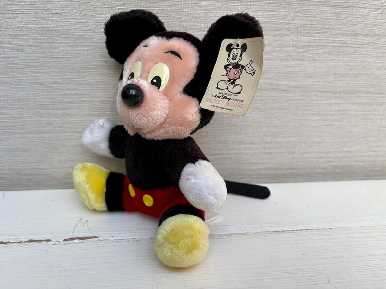 Puede incluir: Un peluche de Mickey Mouse con detalles en negro, rojo, amarillo y blanco. El juguete tiene una cara rosa, orejas negras y una cola negra. Una etiqueta con el logotipo de Mickey Mouse est&aacute; adjunta.