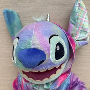 May include: A plush Stitch doll with a colorful tie-dye design. The doll has large, expressive eyes, a blue nose, and a wide grin. The ears are patterned with a plaid design. The doll is wearing a matching hoodie. The overall color scheme includes shades of blue, pink, and purple.