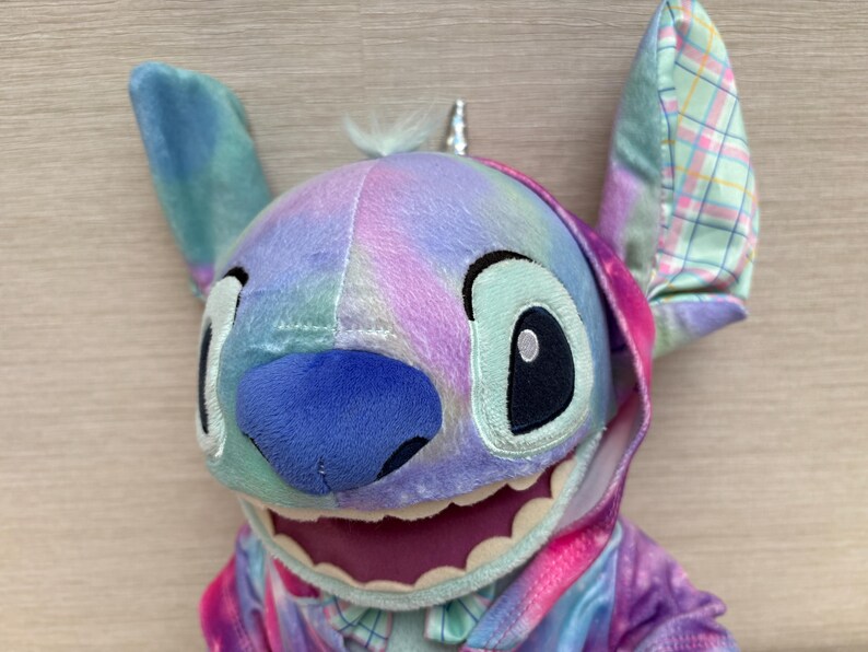 May include: A plush Stitch doll with a colorful tie-dye design. The doll has large, expressive eyes, a blue nose, and a wide grin. The ears are patterned with a plaid design. The doll is wearing a matching hoodie. The overall color scheme includes shades of blue, pink, and purple.