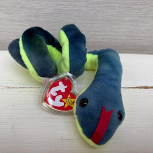 May include: A plush snake toy featuring a dark blue top and a bright lime green underside. The stuffed animal has a red felt tongue and two small black eyes. A heart-shaped red Ty Beanie Baby tag is attached to the side of the coiled body.