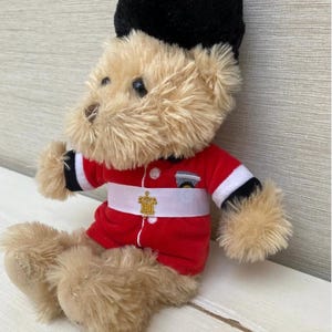 May include: A plush teddy bear dressed as a British Royal Guard. The bear wears a red jacket with white and black trim, a white belt with a gold emblem, and a tall black hat. The bear is sitting on a white surface.