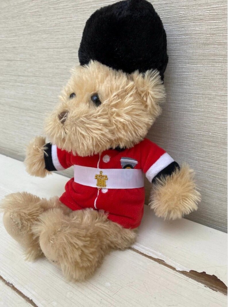 May include: A plush teddy bear dressed as a British Royal Guard. The bear wears a red jacket with white and black trim, a white belt with a gold emblem, and a tall black hat. The bear is sitting on a white surface.
