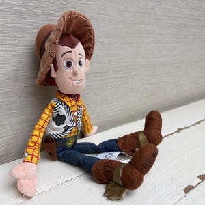 May include: A plush Woody doll from the Toy Story franchise. The doll features a brown cowboy hat, a yellow plaid shirt, a black and white vest, blue jeans, and brown boots. The doll is sitting on a white surface.