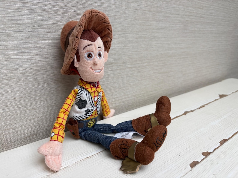 May include: A plush Woody doll from the Toy Story franchise. The doll features a brown cowboy hat, a yellow plaid shirt, a black and white vest, blue jeans, and brown boots. The doll is sitting on a white surface.