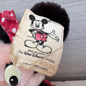 K&ouml;nnte beinhalten: Ein Minnie Mouse Pl&uuml;schtier mit einer rot-wei&szlig; gepunkteten Schleife. Das Etikett zeigt eine klassische Illustration von Mickey Mouse und den Text "MADE EXCLUSIVELY The Walt Disney Company MINNIE MOUSE".