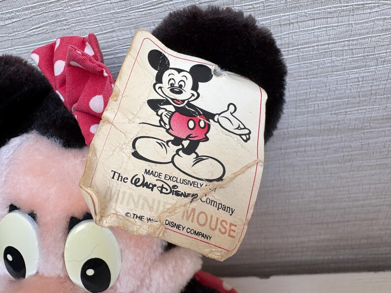 K&ouml;nnte beinhalten: Ein Minnie Mouse Pl&uuml;schtier mit einer rot-wei&szlig; gepunkteten Schleife. Das Etikett zeigt eine klassische Illustration von Mickey Mouse und den Text "MADE EXCLUSIVELY The Walt Disney Company MINNIE MOUSE".