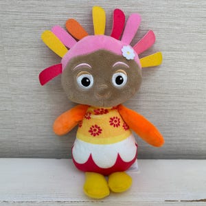 May include: A plush toy character with a brown face, large white eyes, and a pink headband featuring a small white flower. The hair consists of multiple colorful felt strips in yellow, orange, pink, and red. The body has a yellow top with red flower patterns and a white and red scalloped skirt with yellow feet.