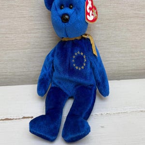 May include: A plush blue teddy bear with a black nose and a gold ribbon around its neck. The bear has a gold emblem of stars on its chest and a red tag attached to its ear.